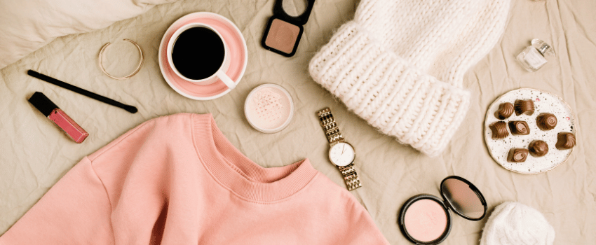 A cozy scene features a pink sweater, a coffee cup, and assorted items neatly placed on a bed, with the sweater bought using teacher clothing discounts.