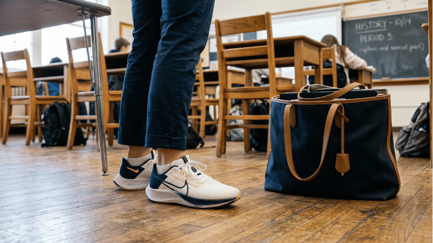Teacher wearing clean athletic sneakers standing next to a large premium tote bag in a classroom.